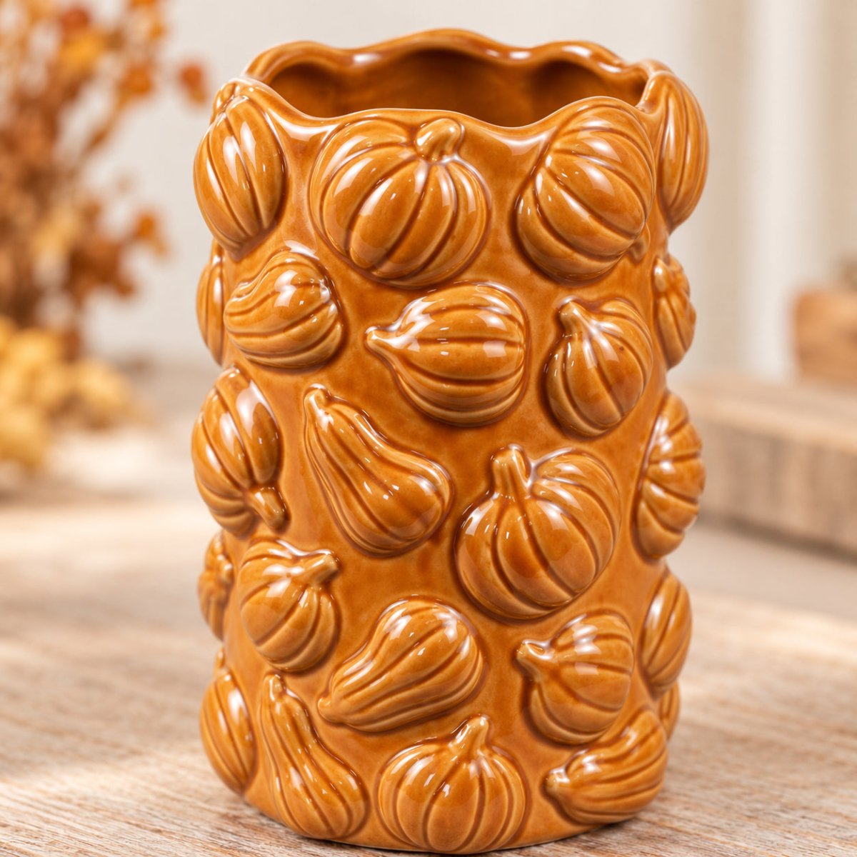 An orange glazed ceramic vase featuring a raised relief pattern of pumpkins, set on a wooden surface with a softly blurred background.