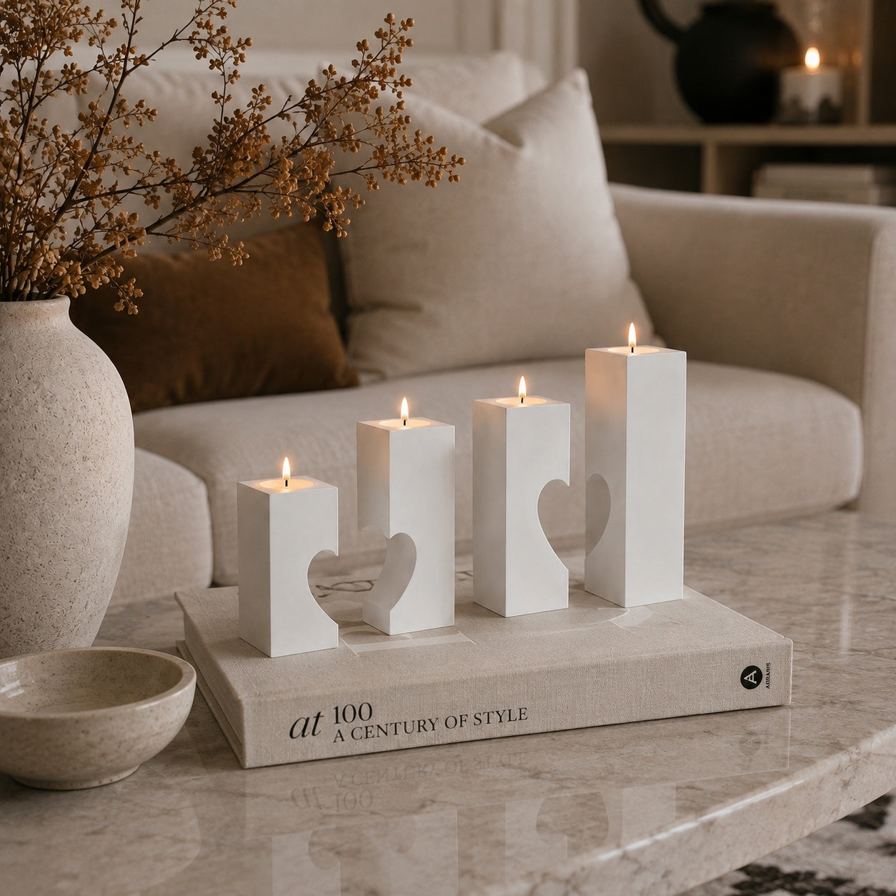 Four white geometric candle holders with heart-shaped cutouts, lit and arranged on a book atop a marble coffee table in a cozy living room setting. styled for modern home decor
