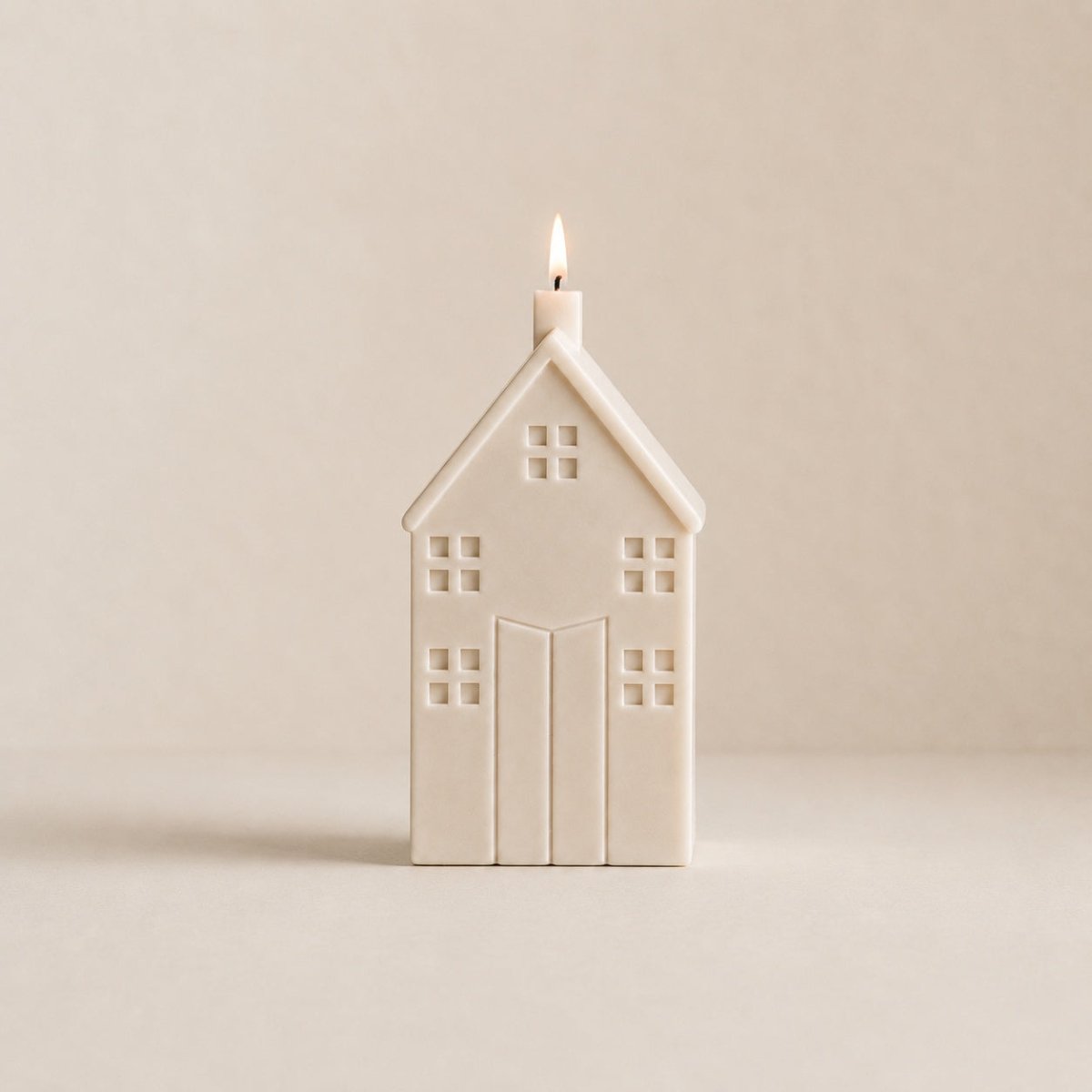 A house-shaped candle with a small flame burning from the top, made of a matte cream wax. The candle is centered against a solid, warm beige background.