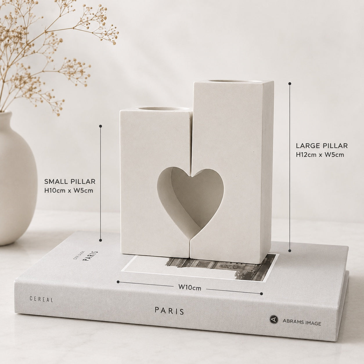 Two minimalist white Handcrafted stone candle holders shaped like pillars, with one featuring a heart-shaped cutout, displayed on top of a book with dimension callouts showing measurements.