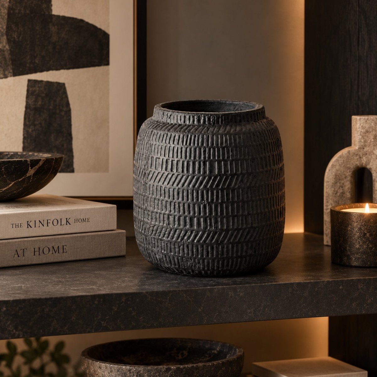A dark textured ceramic vase with a ribbed basketweave pattern, displayed on a dark stone shelf next to books and a lit candle.
