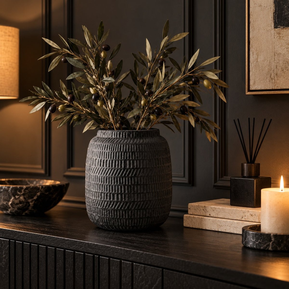A dark textured ceramic vase filled with olive branches and small olives, styled on a dark wooden console table in a moody room with candles, a diffuser, and wall paneling.