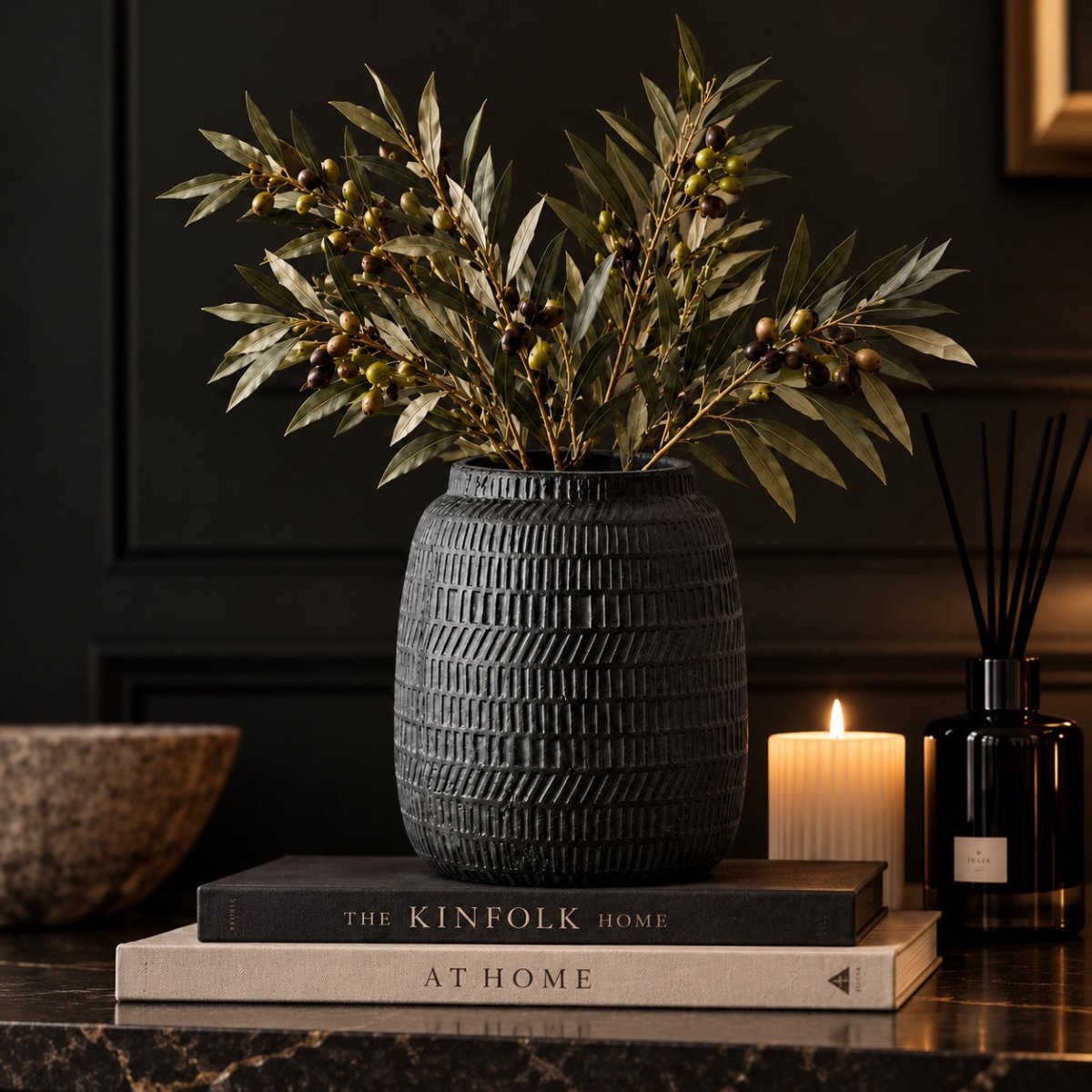 A black textured ceramic vase filled with dried olive branches, styled on a stack of books on a dark marble surface next to a lit candle and a reed diffuser.