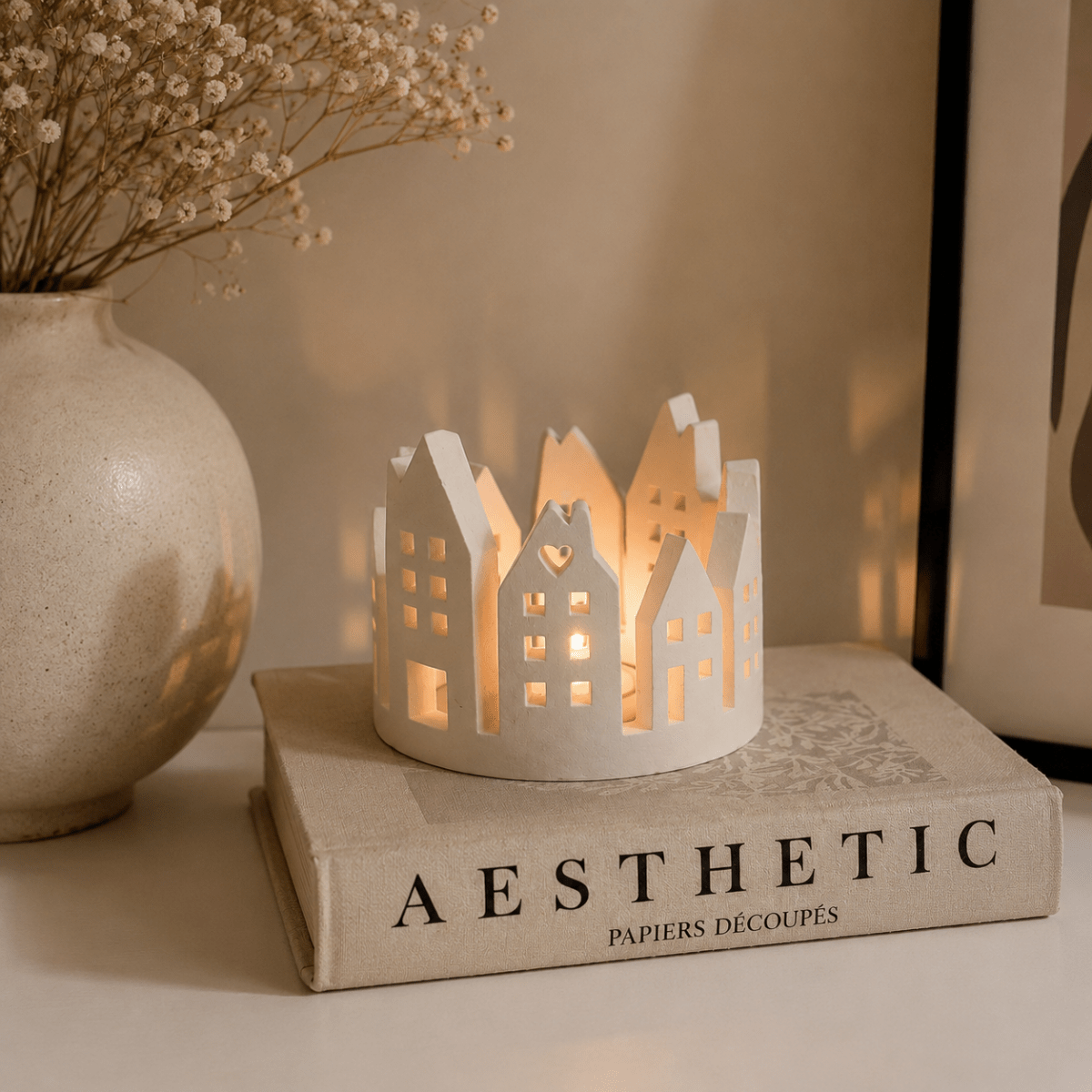 A white handcrafted stone miniature house-shaped tea light candle holder with a warm glowing candle inside, displayed on a decorative book next to a ceramic vase with dried baby's breath. For autumn home styling
