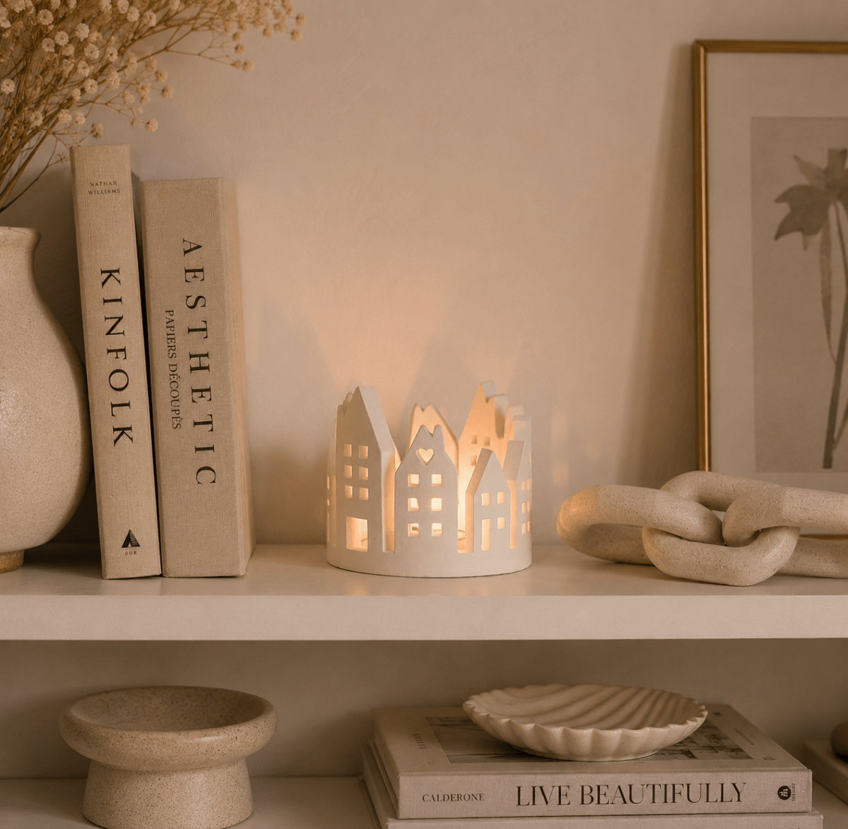 A white stone house-shaped candle holder with glowing warm tealight inside, styled on a shelf next to books, a vase, and decorative objects in a warm, neutral interior setting.