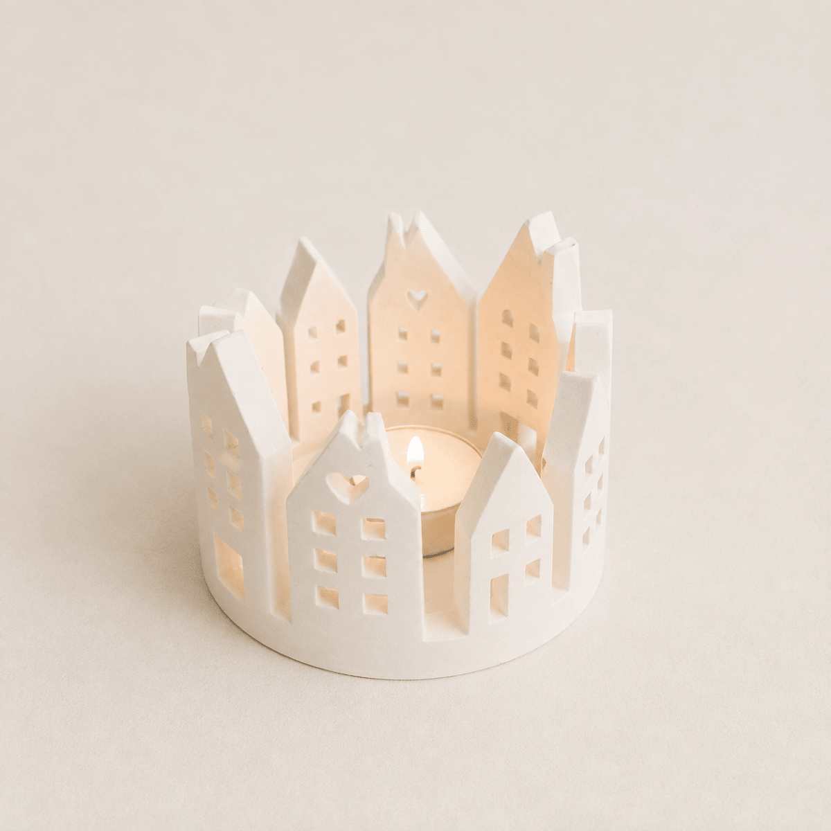 A white handmade stone Dutch gable house candle holder with a lit tea light candle inside, set against a warm beige background.