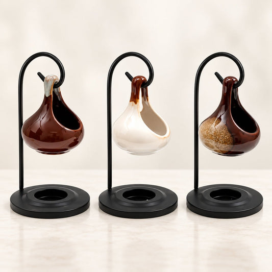 Three small hanging ceramic vessels on black metal stands, arranged side by side on a light marble-like surface against a soft cream background. The vessels have glossy glazes in dark brown, off-white, and a mottled brown-and-beige pattern.