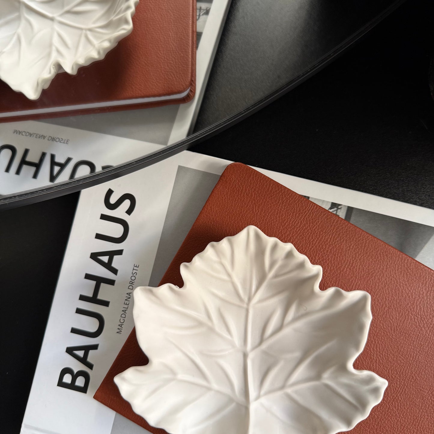 A white Stone leaf-shaped dish with detailed leaf veins, resting on a brown textured notebook over a magazine page with the word 'BAUHAUS' visible.