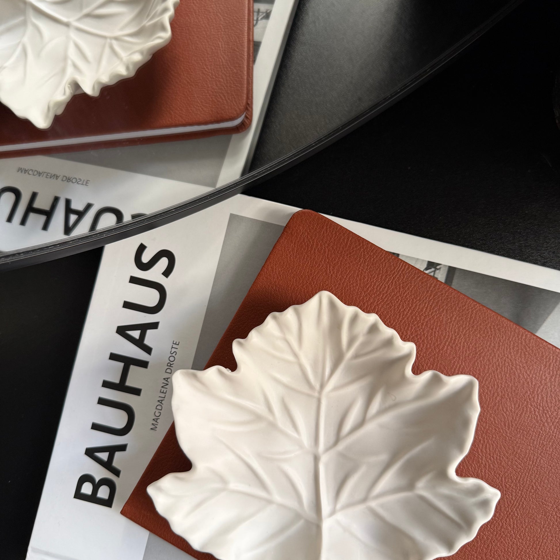 A white Stone leaf-shaped dish with detailed leaf veins, resting on a brown textured notebook over a magazine page with the word 'BAUHAUS' visible.
