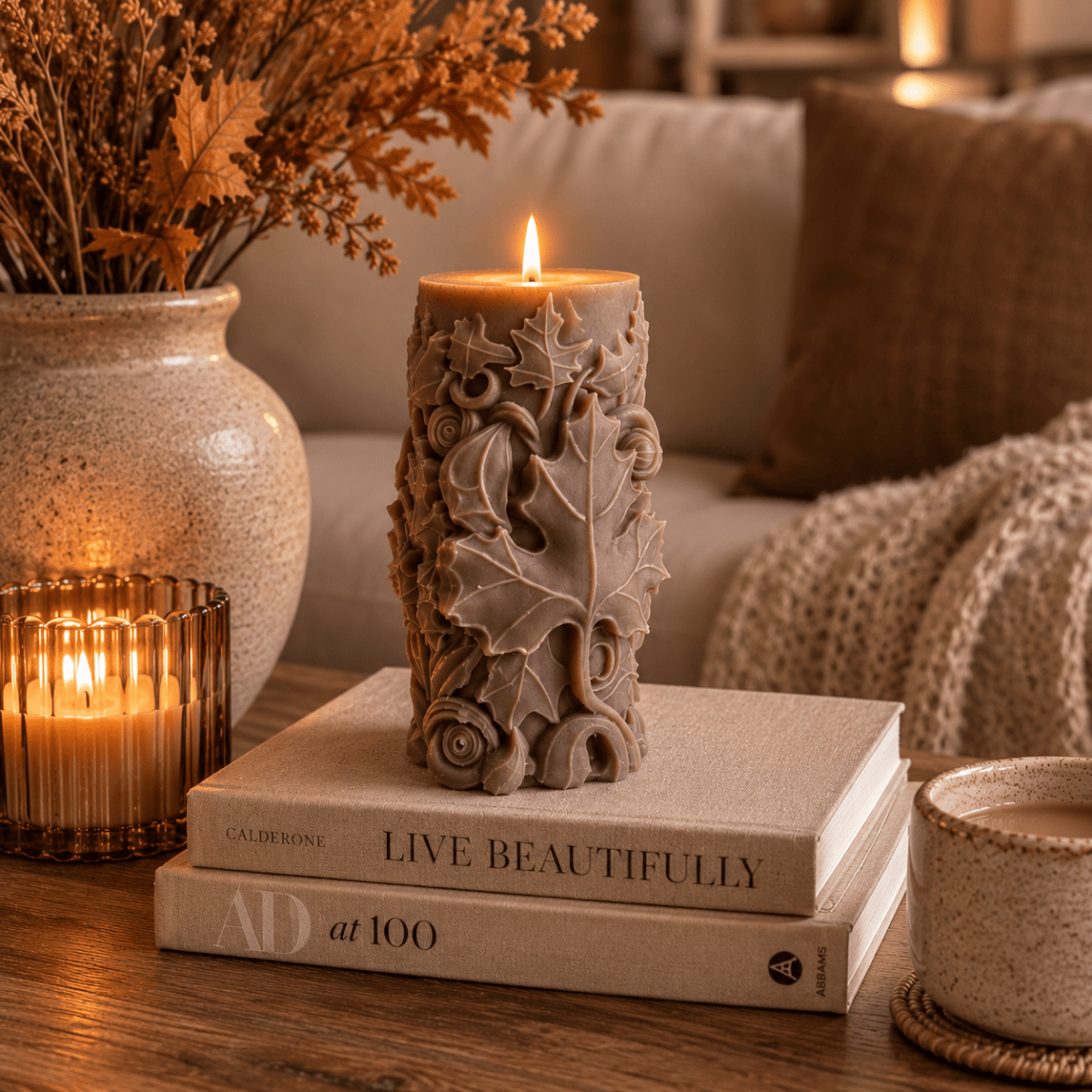 A sculpture pillar candle with a floral and leaf design sits on top of two stacked books on a wooden table, with a lit candle in a ribbed glass holder and a cosy living room background.