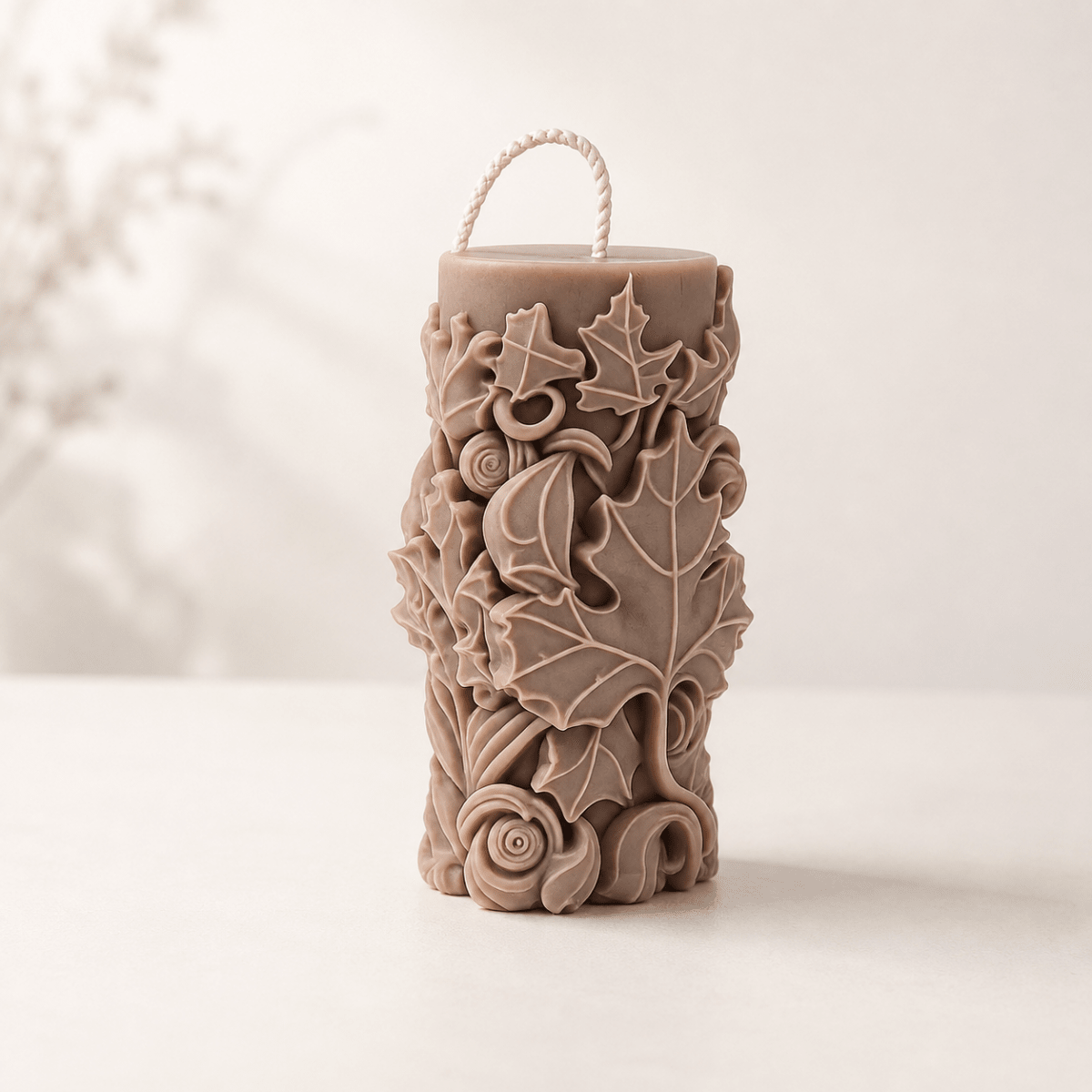 A cylindrical decorative candle, featuring raised floral and maple leaf motifs in a warm taupe-brown colour, set against a soft, light beige background.
