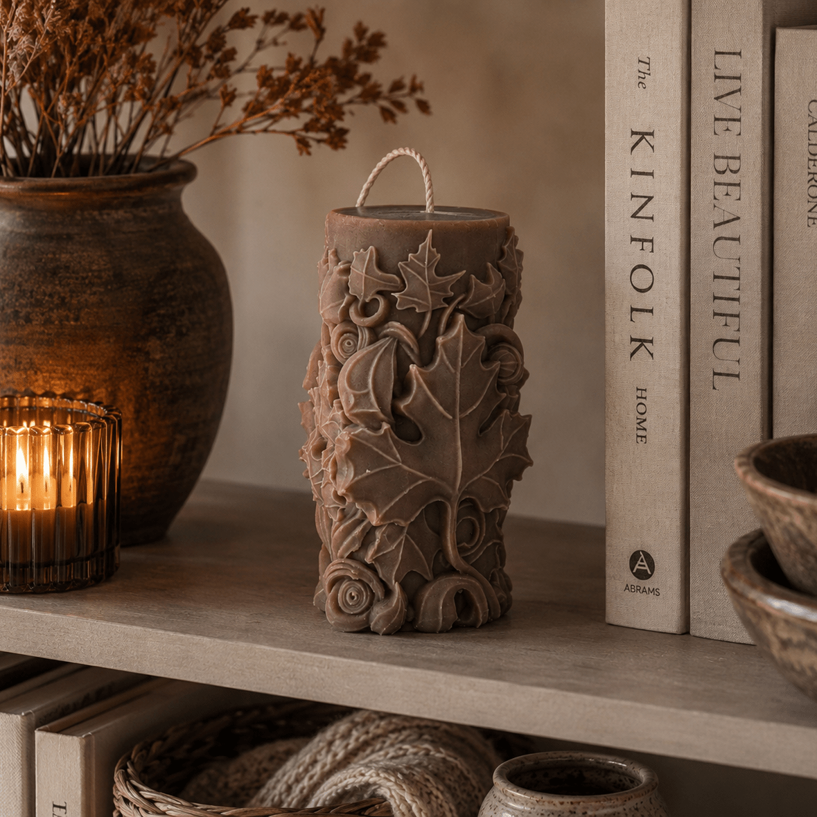 A  brown pillar candle with carved maple leaf patterns sits on a wooden shelf next to a lit candle in a glass holder, books, and ceramic bowls.