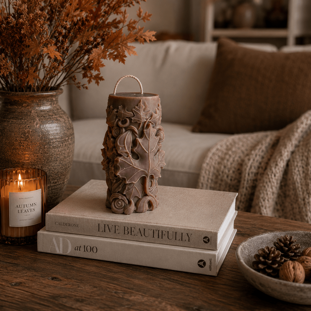 A Sculptured brown soy candle with a leaf and floral pattern sits on a stack of two books on a wooden table, styled in a cosy living room setting with a vase of dried leaves, a lit candle, and a knitted throw blanket in the background.