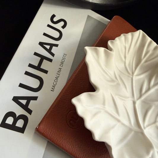 A close-up styled product shot showing a white Handcrafted Jesmonite maple leaf decorative tray resting on a brown textured notebook, with a white book printed with the word BAUHAUS visible underneath.