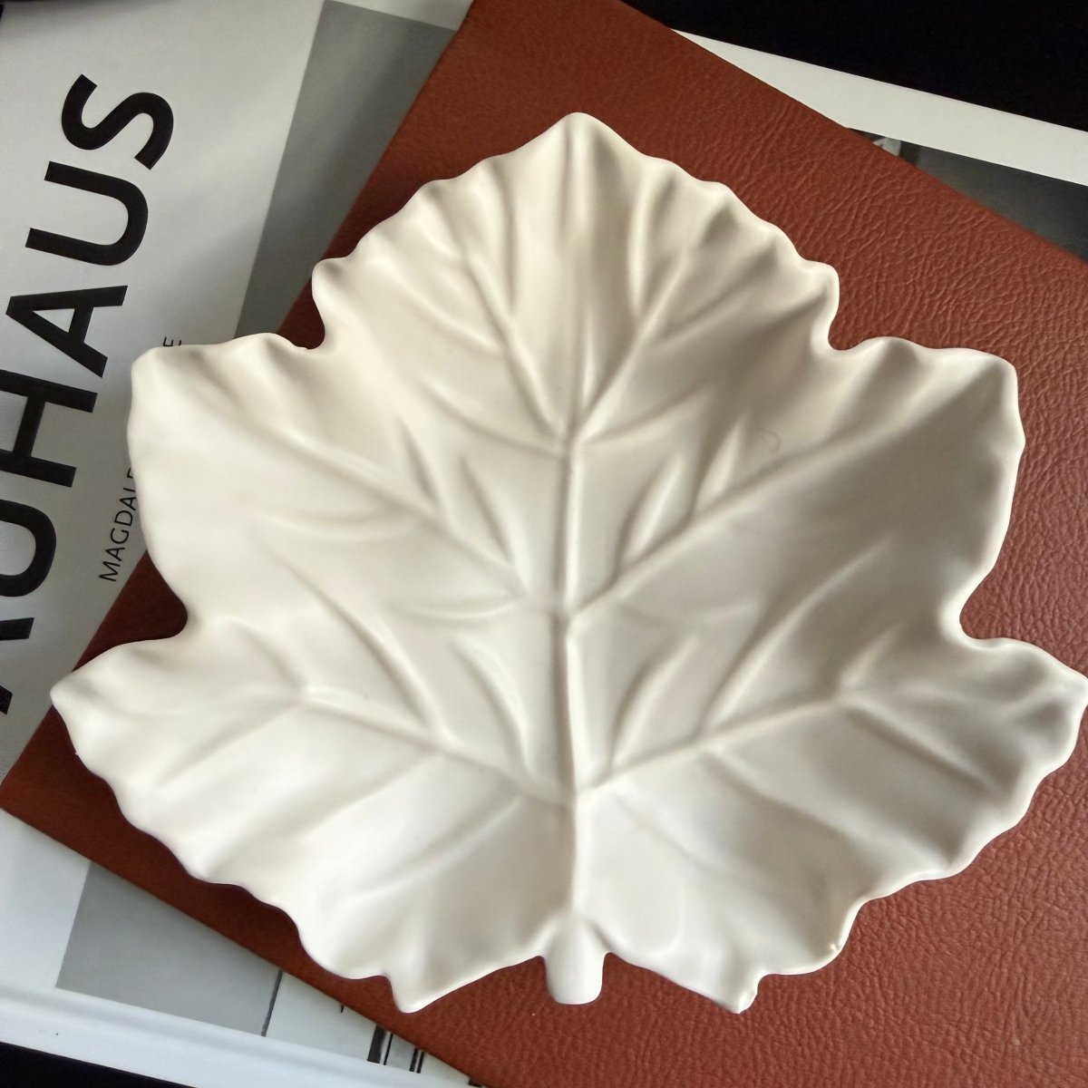 A white leaf-shaped decorative stone tray with detailed embossed leaf veins, resting on a brown textured book or folder over a white magazine page.
