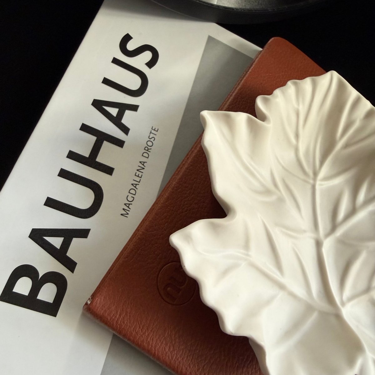 A close-up styled product shot showing a white Handcrafted Jesmonite maple leaf decorative tray resting on a brown textured notebook, with a white book printed with the word BAUHAUS visible underneath.