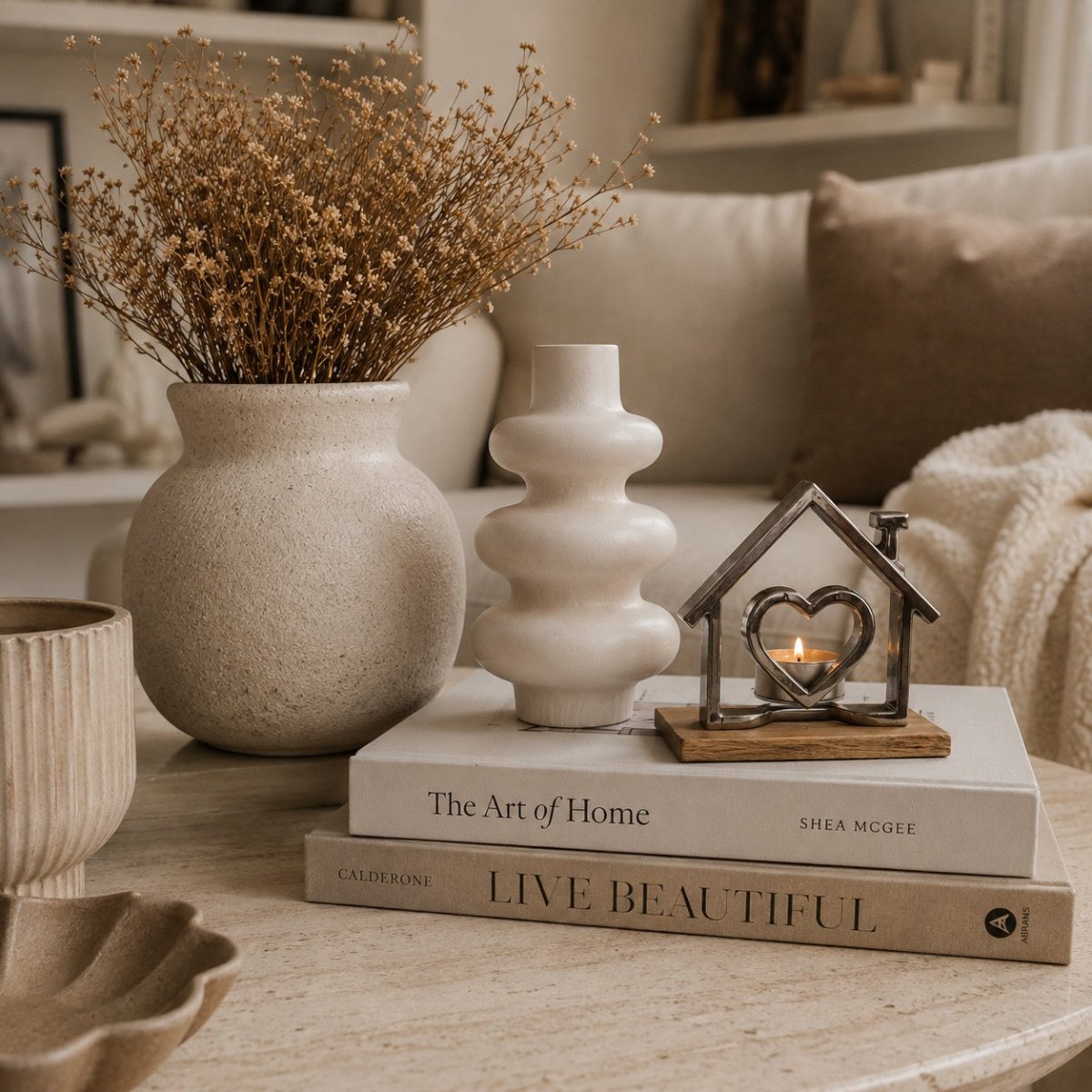 A styled tabletop scene in a cozy living room setting, featuring a textured beige stone vase with dried botanicals, a white sculptural candle holder, a small metal house-shaped candle holder with a heart, and stacked books including 'The Art of Home' and 'LIVE BEAUTIFUL'.