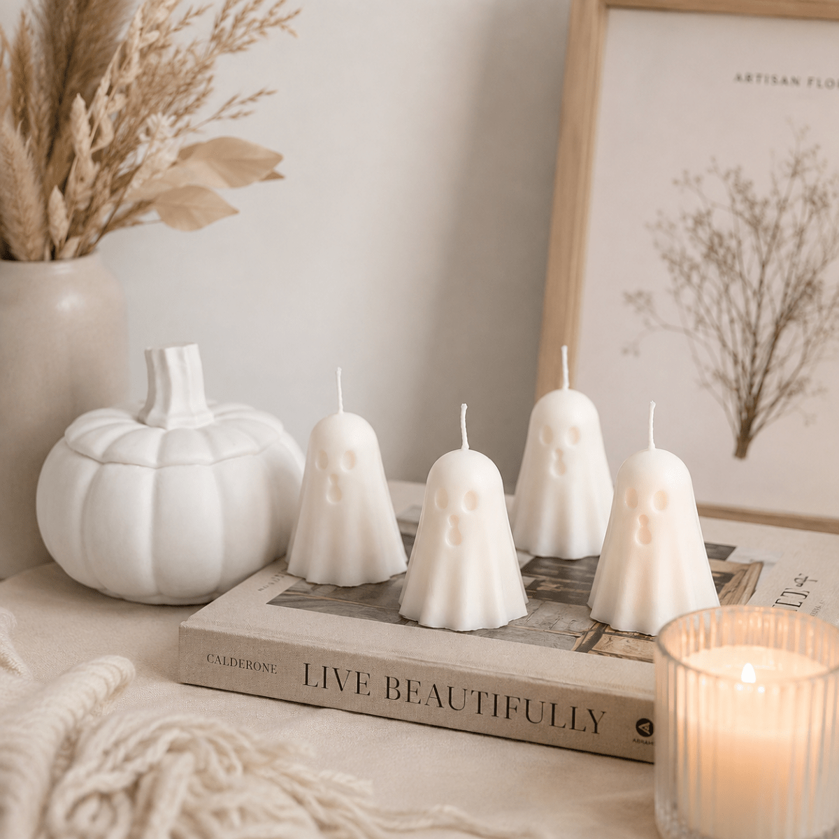 Four white ghost-shaped candles arranged on a book in a styled home setting, with a white pumpkin, dried flowers, a framed print, and a lit candle in the foreground.