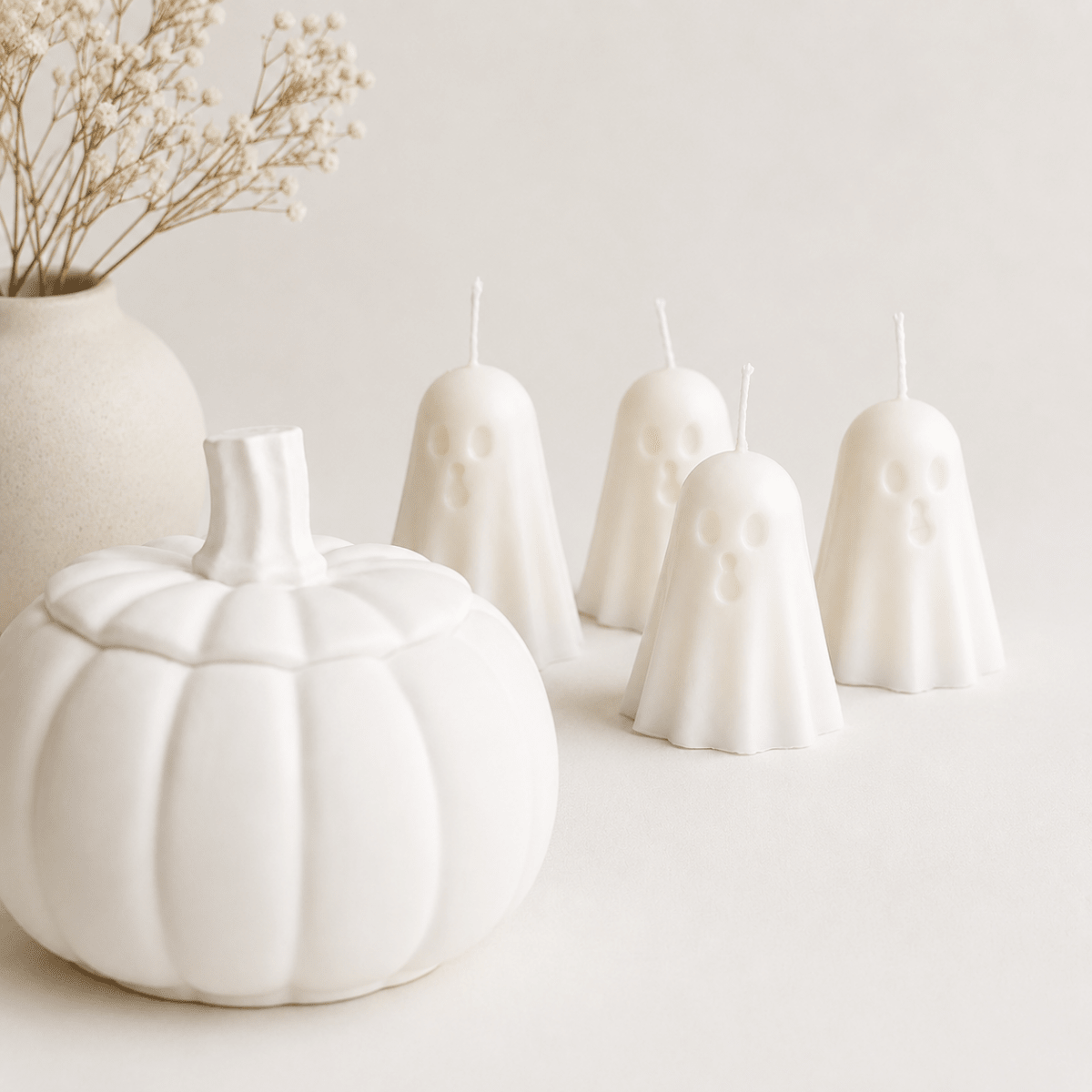 Set of four white ghost-shaped candles styled with a white pumpkin and dried flowers against a light neutral background.