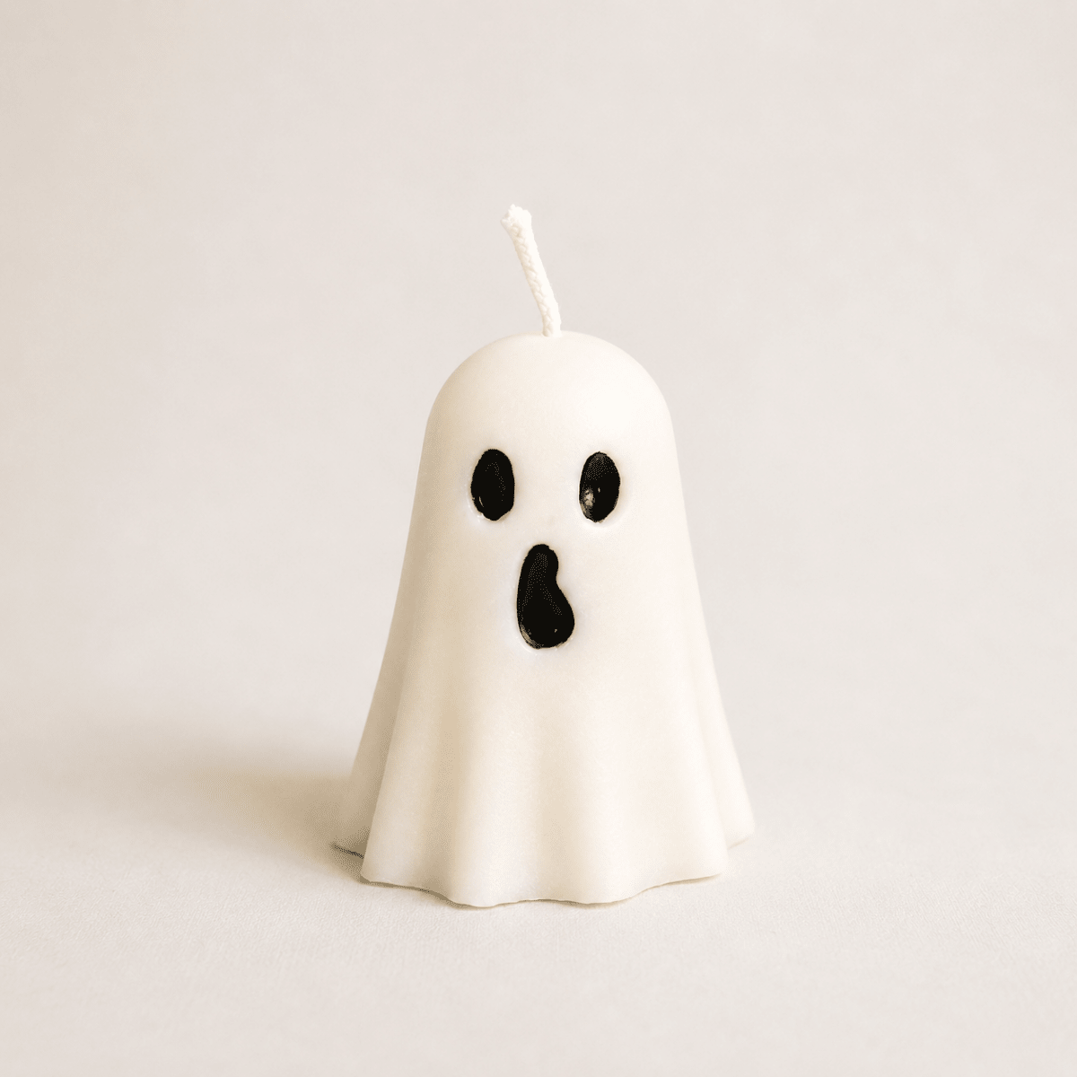 A small ghost-shaped handmade candle in off-white wax with black eyes and an open mouth, standing upright on a light beige background. Autumn home decor