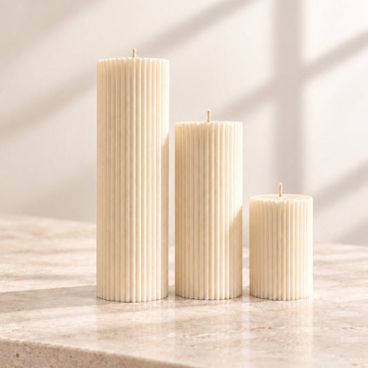 Three ribbed pillar candles in cream-white of different heights standing on a light stone surface with soft window-shadow lighting.