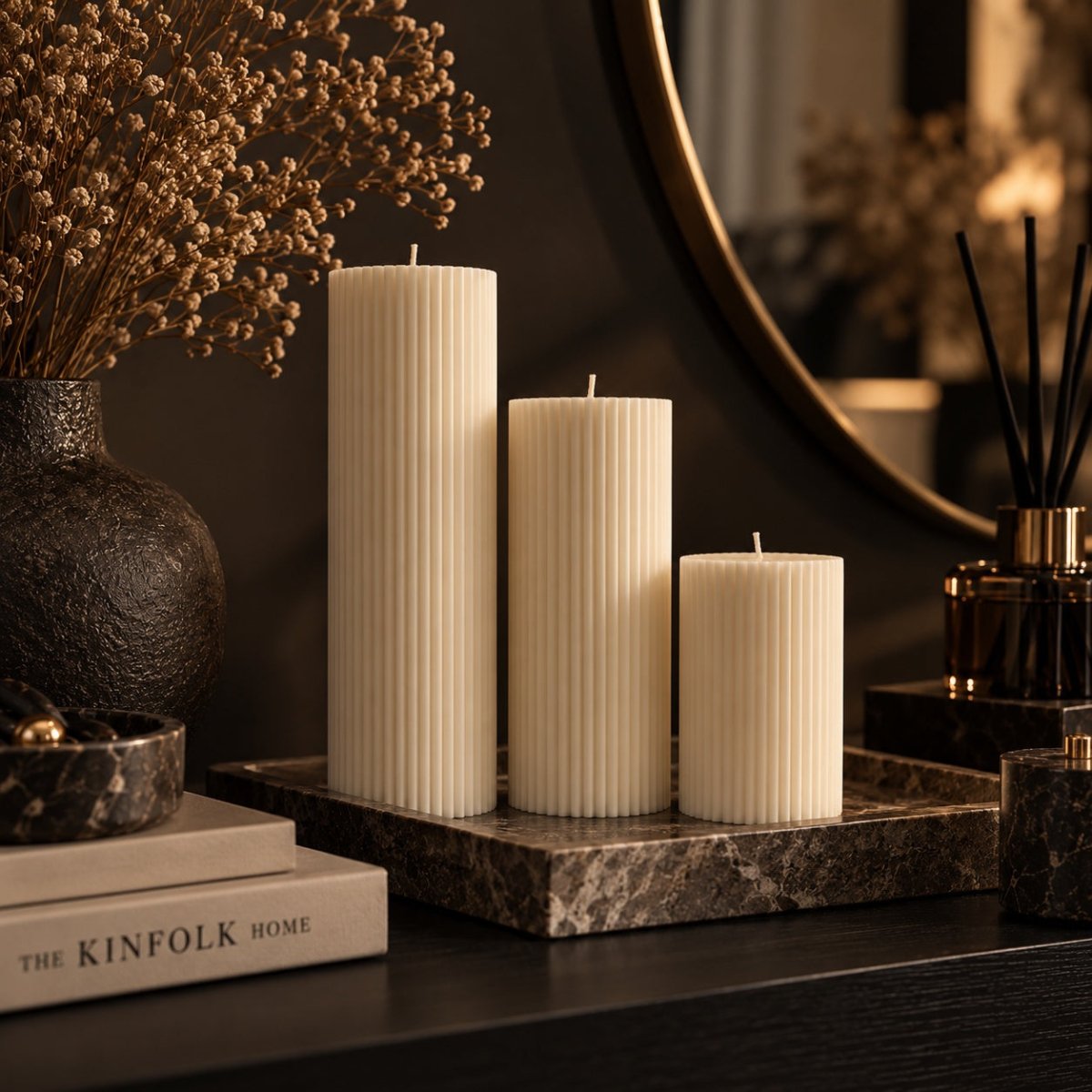 Three cream-colored ribbed pillar candles of varying heights displayed on a dark marble tray on a dark wooden table, styled with dried flowers and home decor.