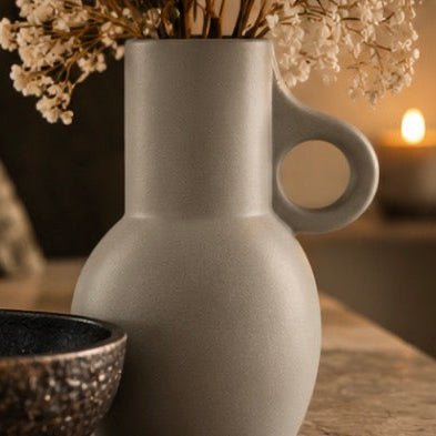 A light grey ceramic vase with a unique side handle, styled with dried white flowers and a dark speckled bowl on a wooden table with a warm candle in the background.