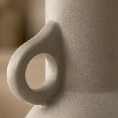 Close-up of a ceramic vessel with a rounded loop handle, shown in warm beige tones against a softly blurred background.