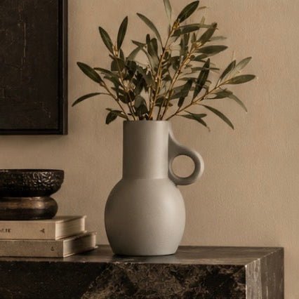 A matte light gray ceramic pitcher vase with a loop handle, holding olive green branches, styled on a dark marble surface against a warm beige wall.