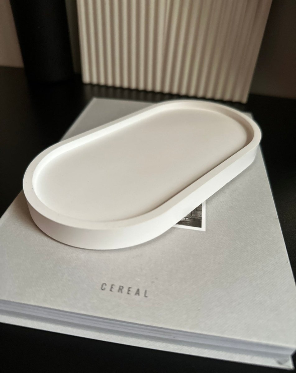 A minimalist, matte white oval tray with a raised rim, resting on a light gray book printed with the word 'CEREAL'. The background features a dark surface and a modern beige ribbed wall panel.