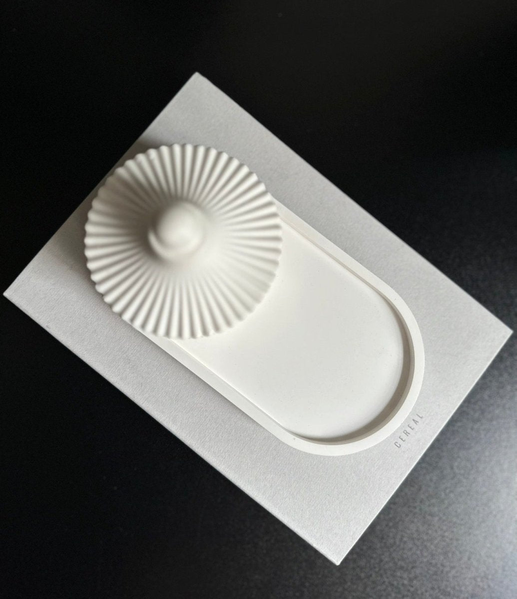 A white ceramic tray with a scalloped circular lid or insert, presented on a textured white backing card against a dark black background.
