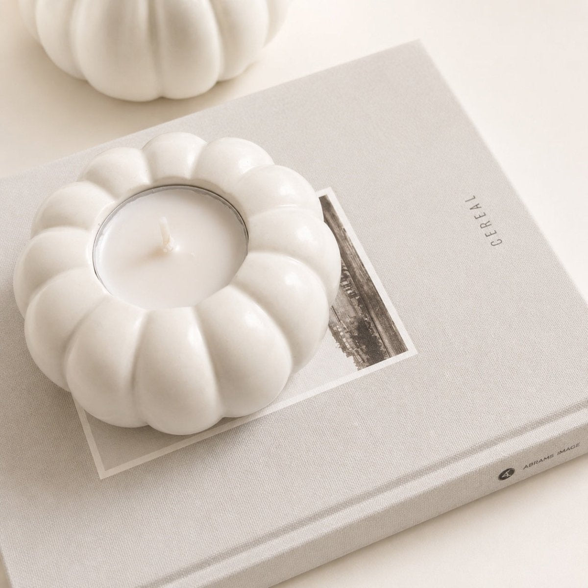 A white handcrafted Stone pumpkin-shaped candle holder containing a white tea light candle, styled on top of a light beige book with a second matching pumpkin candle holder blurred in the background.