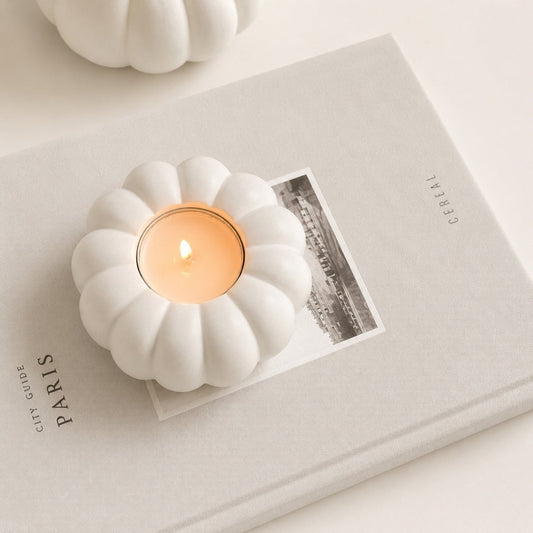 A white stone pumpkin-shaped candle holder containing a lit tealight candle, styled on top of a neutral-toned book with a black-and-white photo insert.