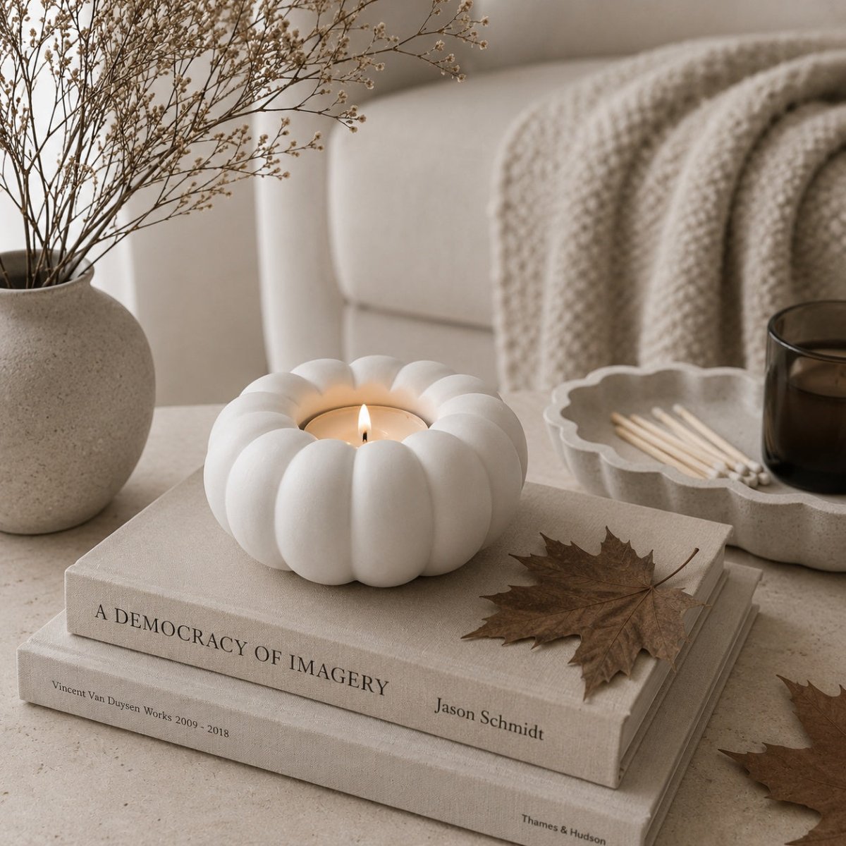 A white pumpkin-shaped candle holder with a lit tealight candle, styled on a stack of books on a light beige surface with dried leaves, a textured throw blanket, and a small vase in the background.