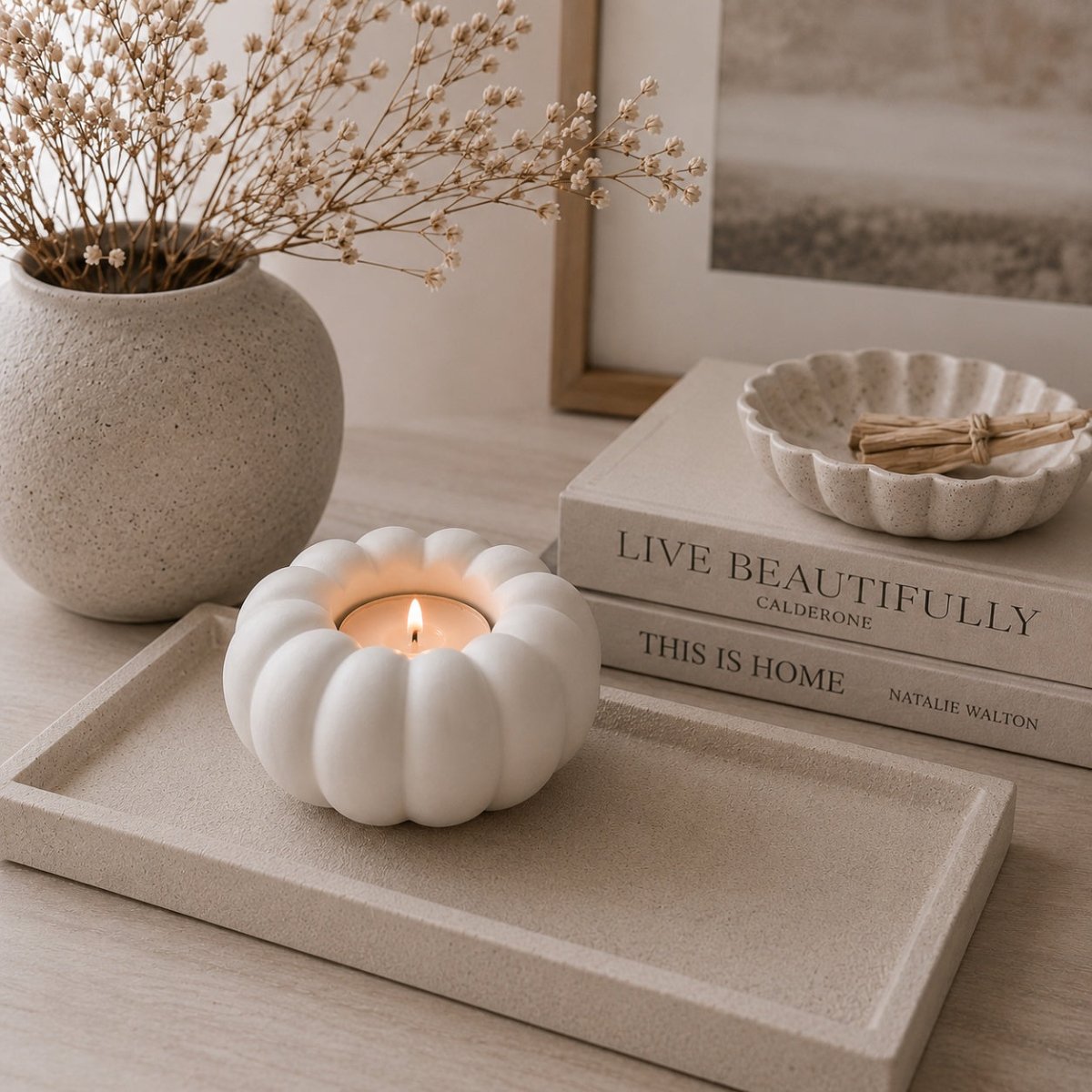 A white ribbed stone tea light candle holder with a lit candle, styled on a beige tray next to a textured vase with dried flowers and stacked books.