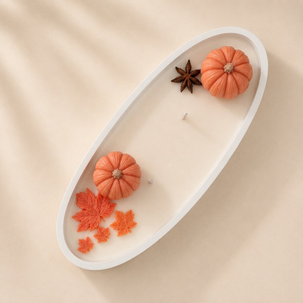 An oval white handmade candle tin with a white wick, styled with miniature orange pumpkins, orange maple leaves, and a brown star anise on a beige fabric background.