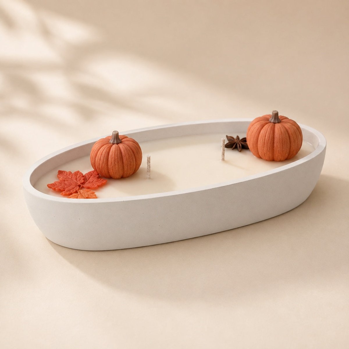 An oval white Stone candle holder filled with cream wax, styled with two small orange pumpkins, a red-orange leaf, and a star anise on a warm beige background.