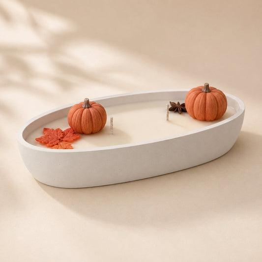 An oval white Stone candle holder filled with cream wax, styled with two small orange pumpkins, a red-orange leaf, and a star anise on a warm beige background.