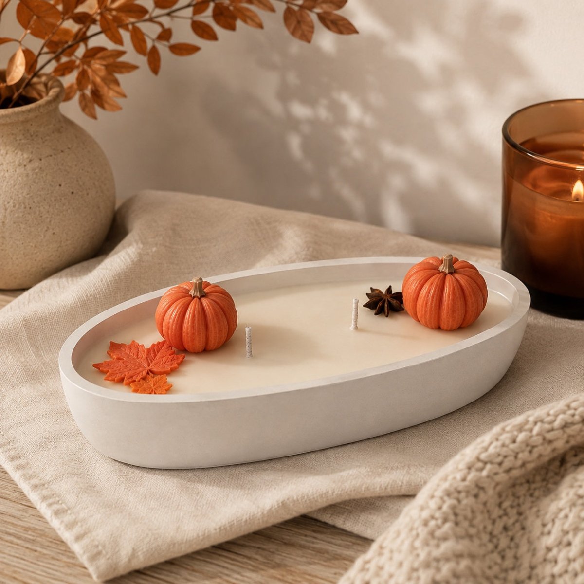 An oval white candle dish holding a cream-coloured candle, styled with two small orange pumpkins, a dried orange leaf, and a star anise, set on a beige cloth with a lit amber candle and a vase of leaves in the background.