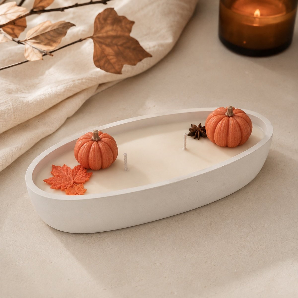 A white oval candle dish or tray holding a white candle, styled with two small orange pumpkins, an orange leaf, and a star anise, on a beige surface with a linen cloth and a lit amber candle in the background.