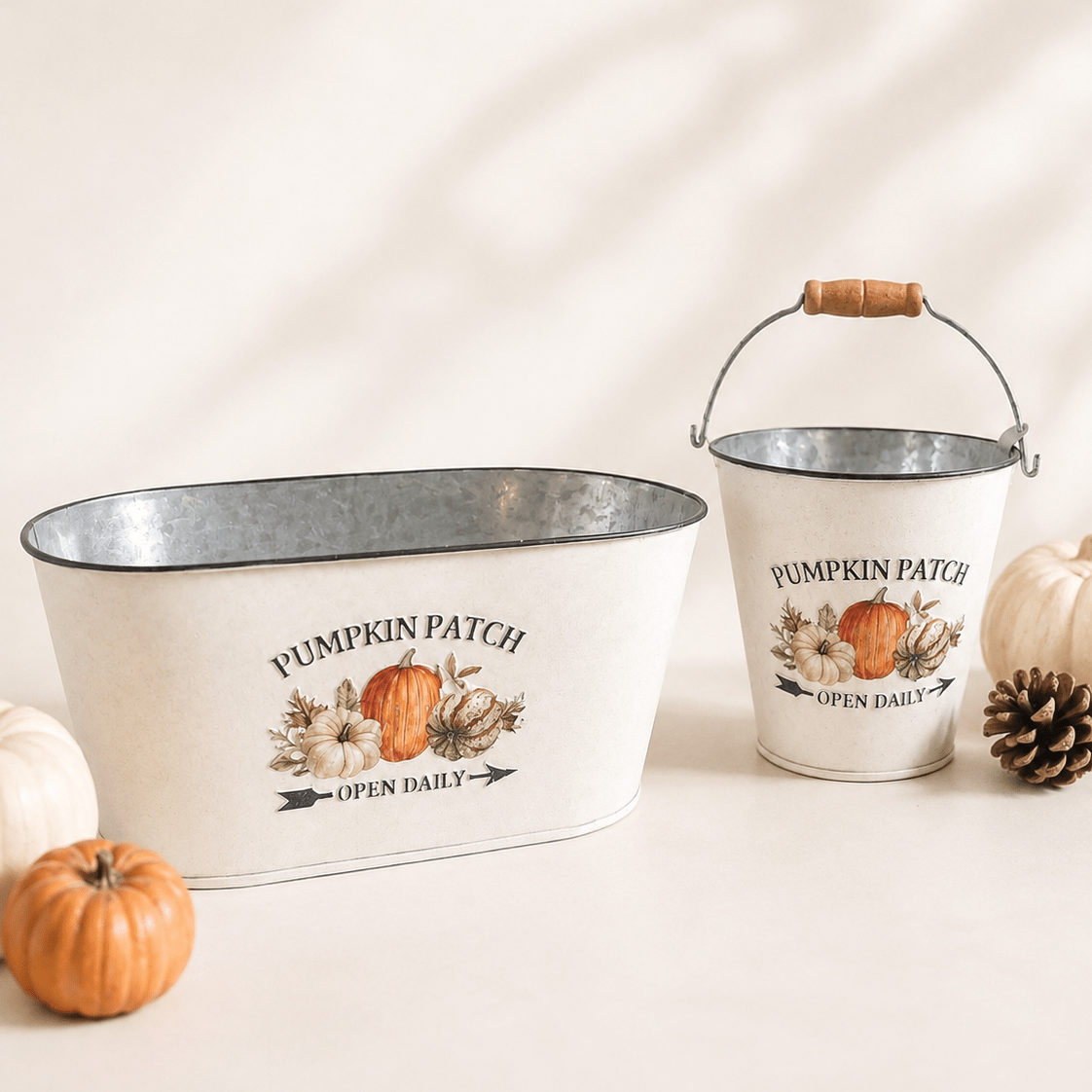 Two decorative Zinc metal buckets with a Autumn Pumpkin theme, one large oval tub and one smaller bucket with a handle, both printed with 'PUMPKIN PATCH OPEN DAILY' and surrounded by small pumpkins, a pinecone, and white pumpkins on a beige background.