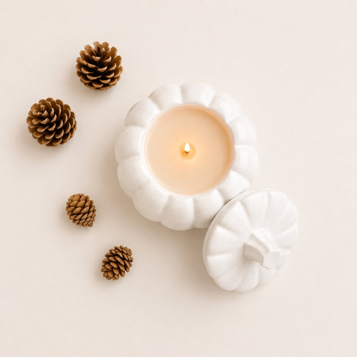 A white stone pumpkin-shaped candle with its lid placed beside it on a warm cream background, styled with four brown pinecones. The candle is lit, showing a soft cream-colored wax and a small glowing flame.
