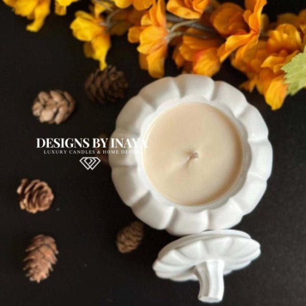 White candle in a decorative holder with yellow flowers and pine cones on a dark background, featuring 'Designs by Inaya'.