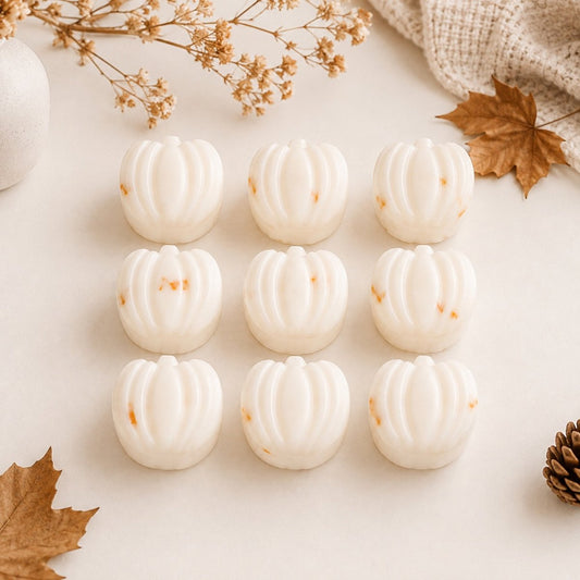 Nine small pumpkin-shaped soy wax melts or soap bars arranged in a 3-by-3 grid on a light cream surface, with autumn-themed props including dried leaves, a pinecone, and a knitted fabric corner.