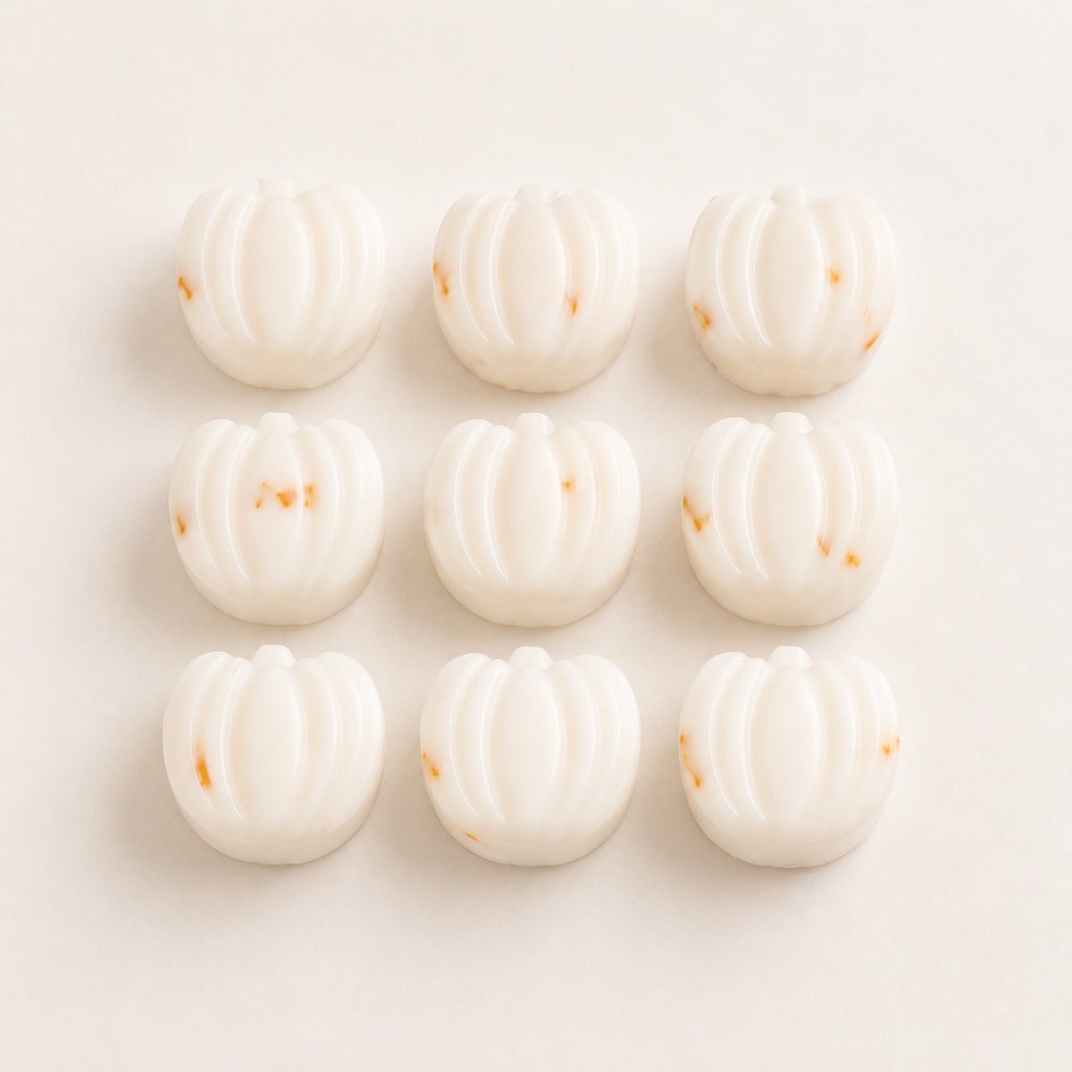 Nine small pumpkin-shaped items arranged in a 3-by-3 grid on a plain light background. The items appear to be matte off-white or cream with a few tiny warm orange specks.
