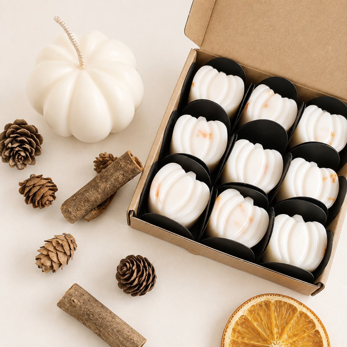 A box of white pumpkin-shaped wax melts or scented bars in black holders, styled on a light beige surface with a white pumpkin, pinecones, cinnamon sticks, and a dried orange slice.
