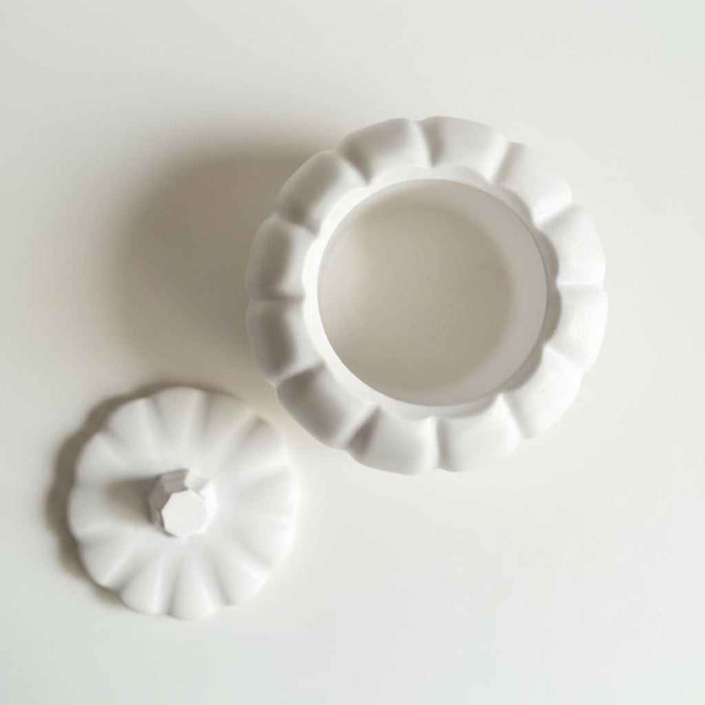 A white handmade stone pumpkin-shaped lidded container with its lid placed beside it on a white surface, styled with pinecones and orange flowers.