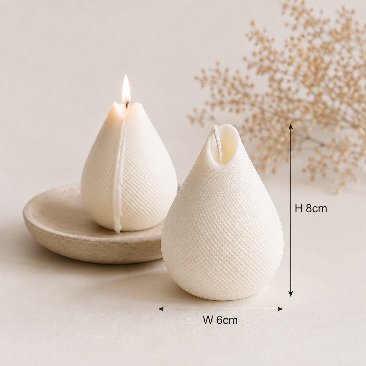 Two textured cream-colored teardrop-shaped soy wax candles on a light beige surface, one lit and resting on a small round dish, with dimension arrows showing height 8cm and width 6cm. A dried floral stem decorates the background.