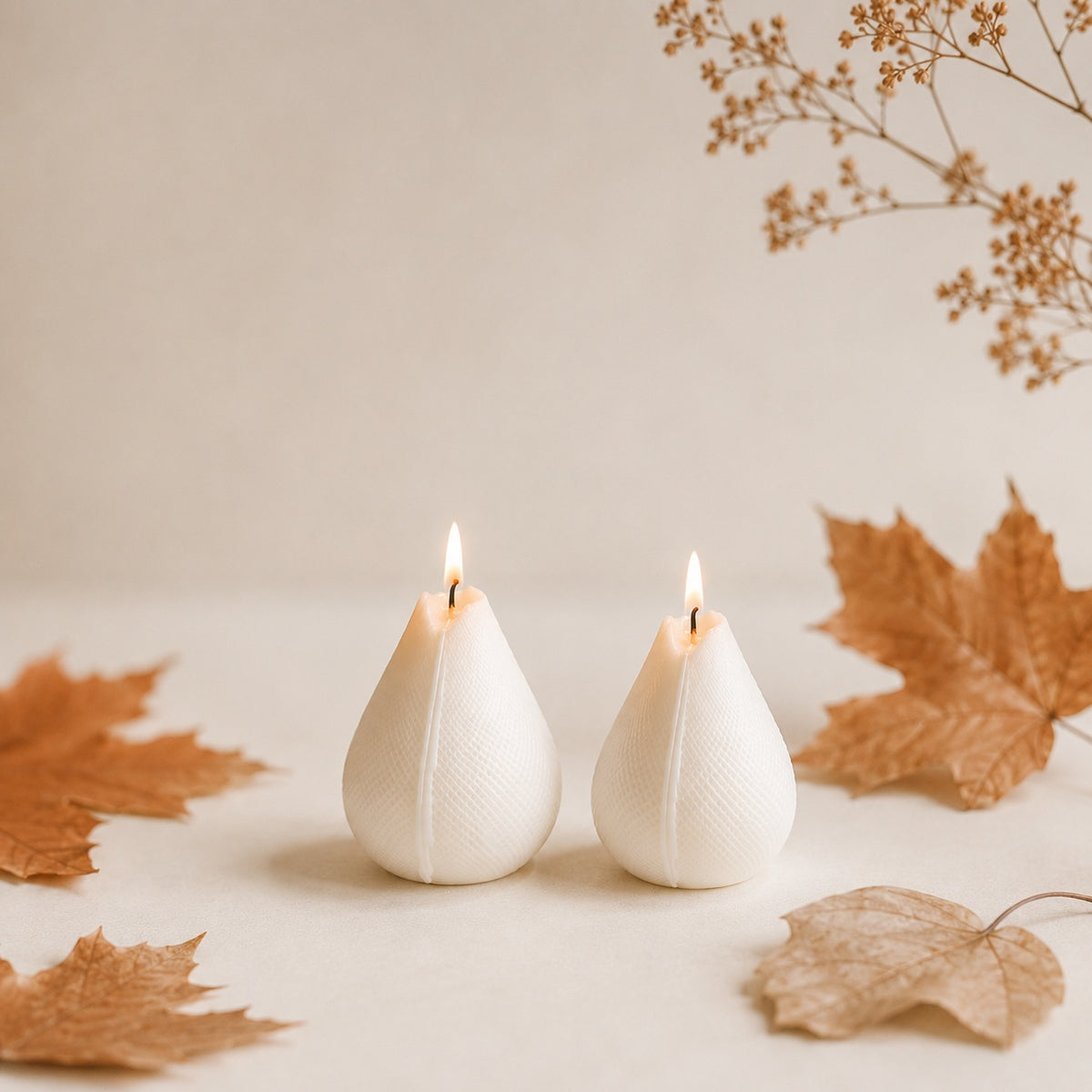 Two cream teardrop-shaped handmade candles with lit flames, styled on a neutral beige surface with scattered dried autumn leaves and a soft beige background.