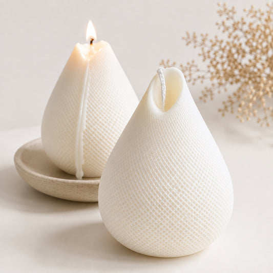 Two white teardrop-shaped textured sculpture candles on a light surface, with one lit and one unlit, styled with dried floral stems in the background.